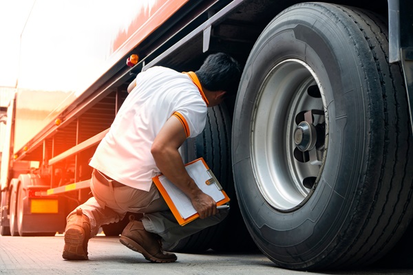 A man crouching down and inspecting the tires of a large truck, holding a clipboard, likely performing a vehicle safety inspection.